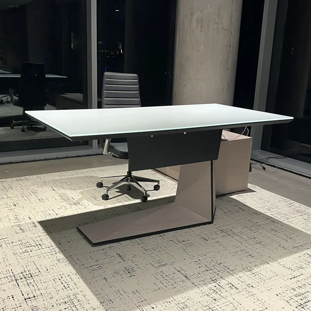 A sleek, modern executive desk with a white top and a unique angled leg design, set over a textured rug with a gray leather desk chair behind it.