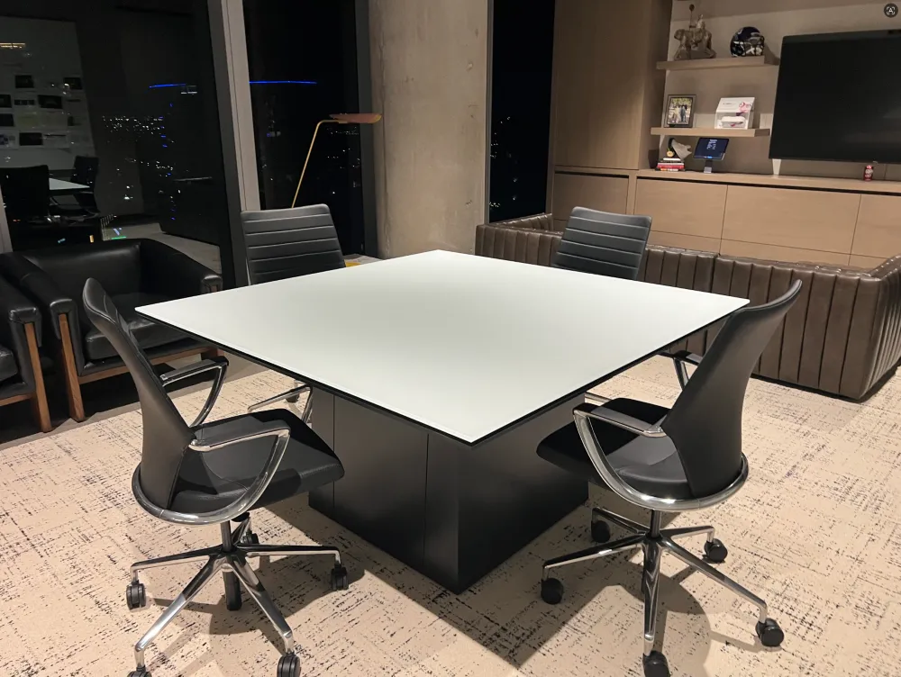 A square white-topped conference table centered in an executive room with four black leather swivel chairs, a brown leather tufted couch, and a wall-mounted TV in the background.