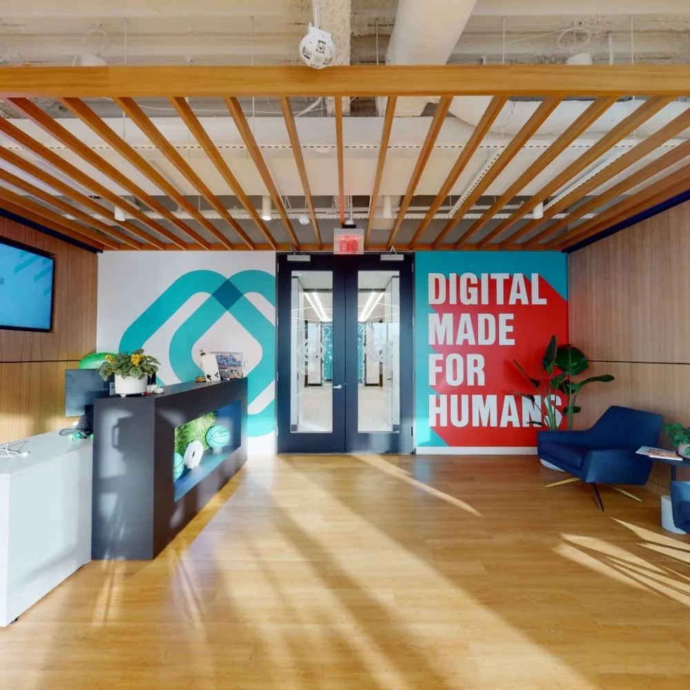 A modern office reception area featuring a black reception desk, warm wood-slatted ceiling beams, and a bold blue and red wall graphic reading "DIGITAL MADE FOR HUMANS".