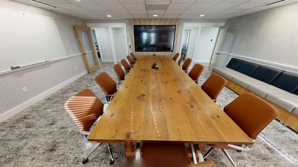 A long natural wood conference table lined with warm brown leather office chairs, with a white wall-mounted board on the left and a media screen at the far end.