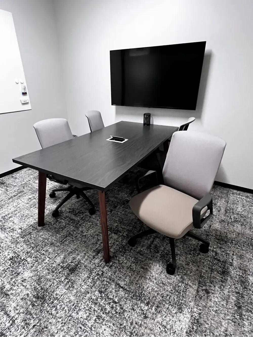 A small meeting space containing a dark wood table paired with light gray office chairs, a wall-mounted TV display, and a textured gray patterned rug.