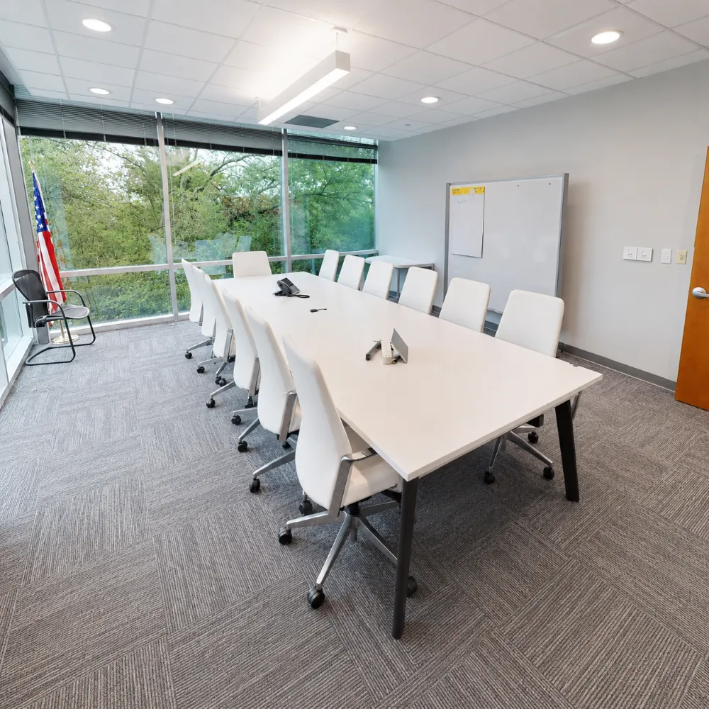 A bright conference room featuring a long white table surrounded by white ergonomic chairs, set against floor-to-ceiling windows overlooking green trees, with an American flag standing in the corner.