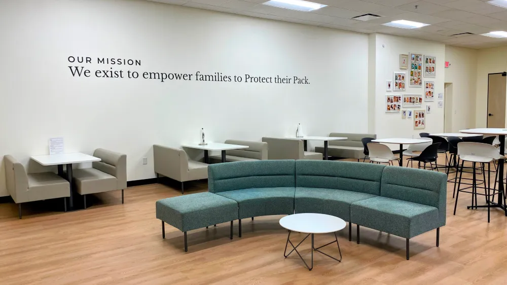 A spacious break area with light wood flooring, featuring a large curved teal sectional sofa, small dining booths, high-top tables, and a wall displaying the quote "OUR MISSION We exist to empower families to Protect their Pack."