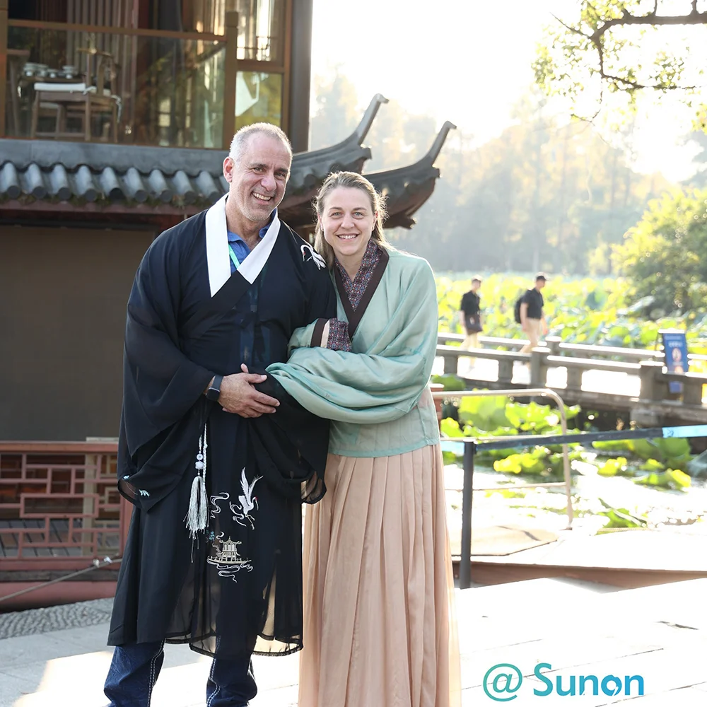 A man and woman wearing traditional East Asian robes posing together outdoors near a scenic water garden and traditional pavilion.