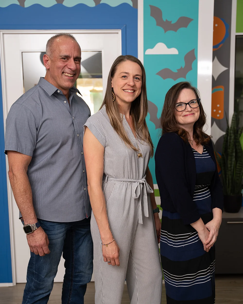 A portrait of three colleagues standing side-by-side in front of a colorful decorated wall, smiling at the camera.