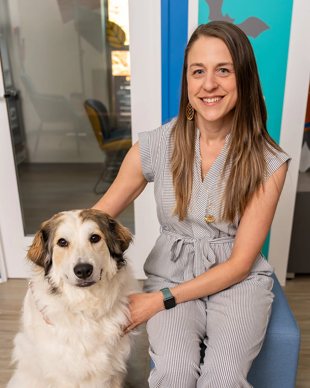 A woman in a striped jumpsuit sitting next to a large, fluffy white and brown dog in an office setting.