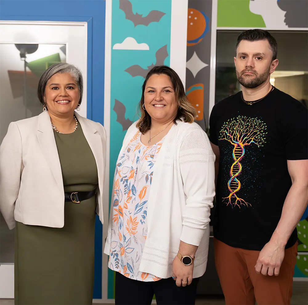 A group portrait of three colleagues standing in front of a colorful decorated wall, with a woman in a green dress on the left, a woman in a white cardigan in the center, and a man in a graphic t-shirt on the right.