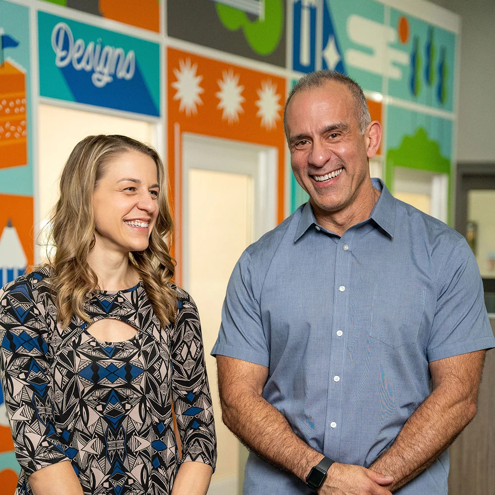 A smiling man and woman standing side-by-side in front of a colorful office wall mural.