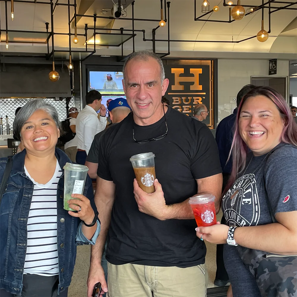 A man in a black t-shirt and two women standing together indoors, smiling while holding Starbucks iced drinks.
